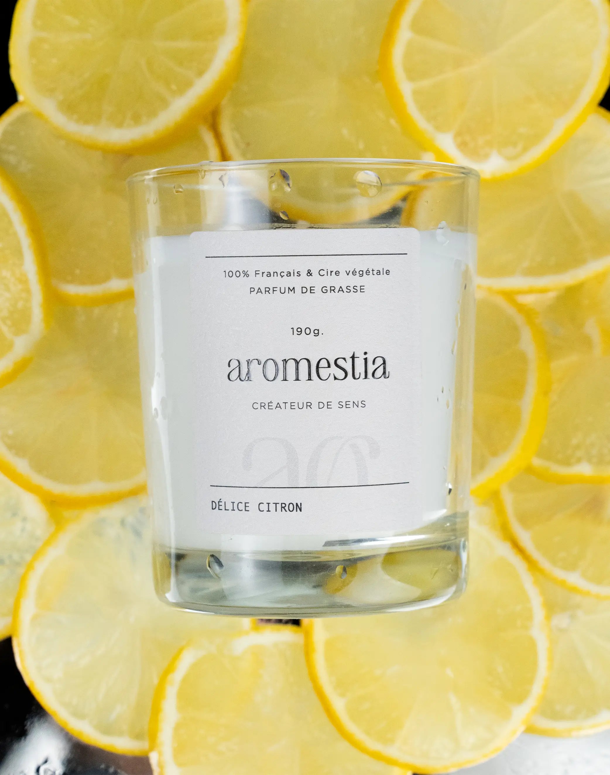 Bougie parfumée délice citron Aromestia avec un contenant transparent et des citrons en arrière-plan